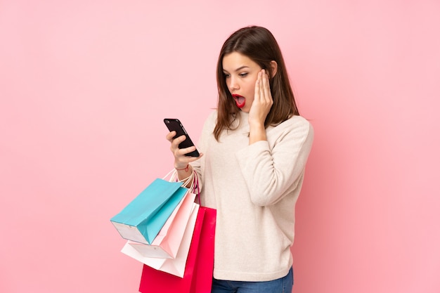 Teenager girl isolated on pink wall holding shopping bags and writing a message with her cell phone to a friend