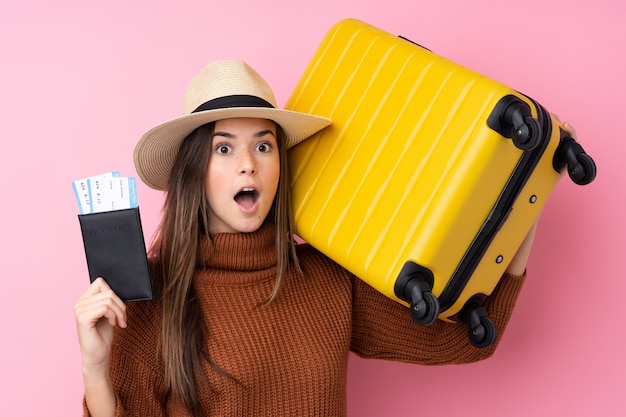 Девушка подростка над изолированной розовой стеной в каникулах с чемоданом и паспортом и удивленная