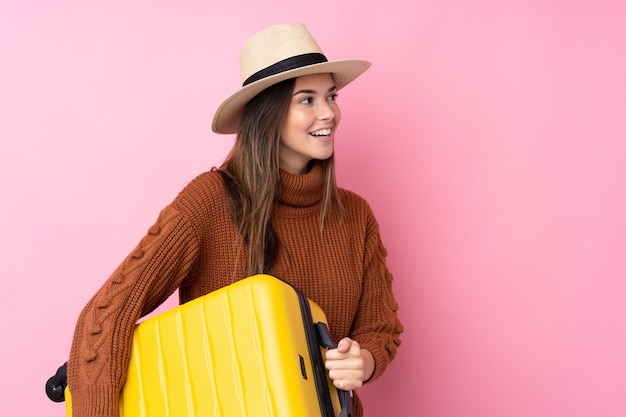 Девушка подростка над изолированной розовой стеной в каникулах с чемоданом перемещения и шляпой