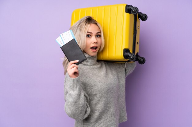 Teenager girl over isolated purple wall in vacation with suitcase and passport and surprised