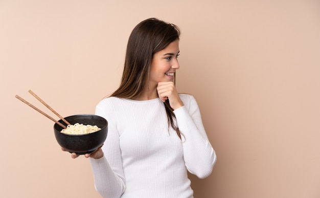 Teenager girl over isolated wall thinking an idea and looking side while holding a bowl of noodles with chopsticks