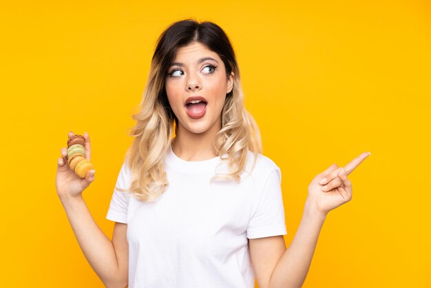 Teenager girl isolated on yellow wall holding colorful French macarons and pointing side