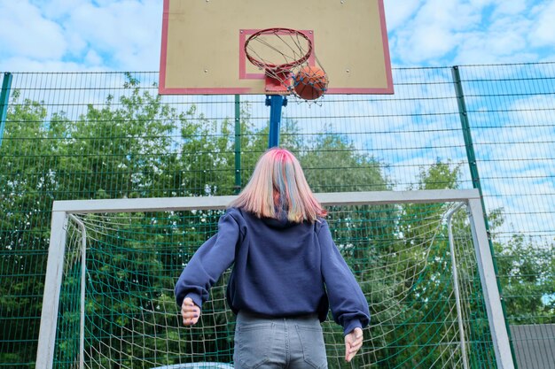 Девушка-подросток прыгает с мячом, играя в уличный баскетбол, вид сзади. Активный здоровый образ жизни, хобби и досуг, молодежная концепция