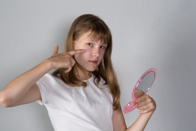 A teenager girl looks at herself in a mirror and looks up a\
pimple