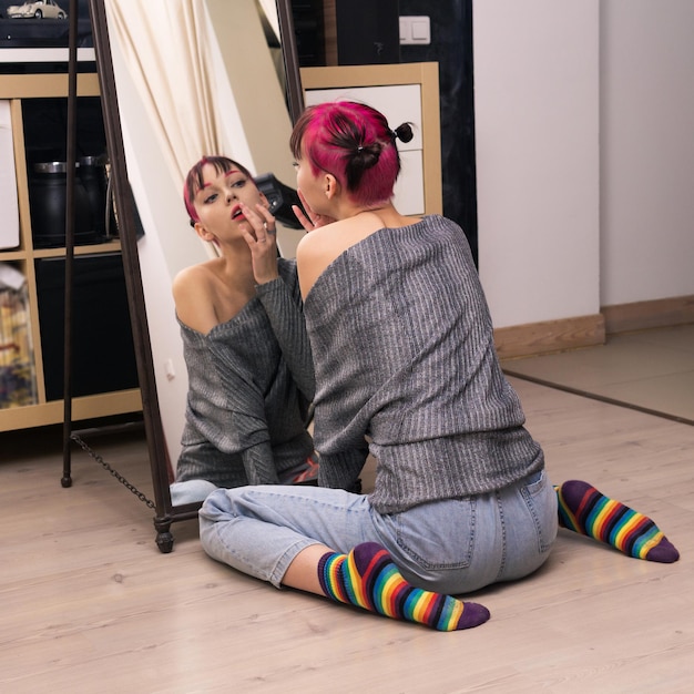 Teenager girl looks in the mirror indoor sitting on the floor carefully examines her face