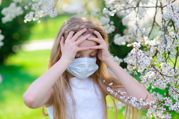 春の花の咲く庭園で医療マスクのティーンエイジャーの女の子。社会的距離の概念とコロナウイルスの予防。