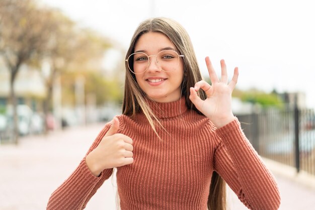 Photo teenager girl at outdoors with glasses and doing ok sign