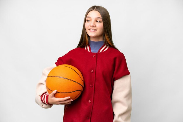 Teenager girl playing basketball over isolated white background looking to the side and smiling