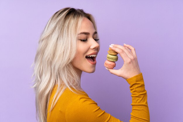 カラフルなフランスのマカロンを押しながらそれを食べる紫色の壁の上のティーンエイジャーの女の子