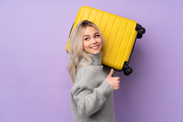 Teenager girl over purple wall in vacation with travel suitcase and with thumb up