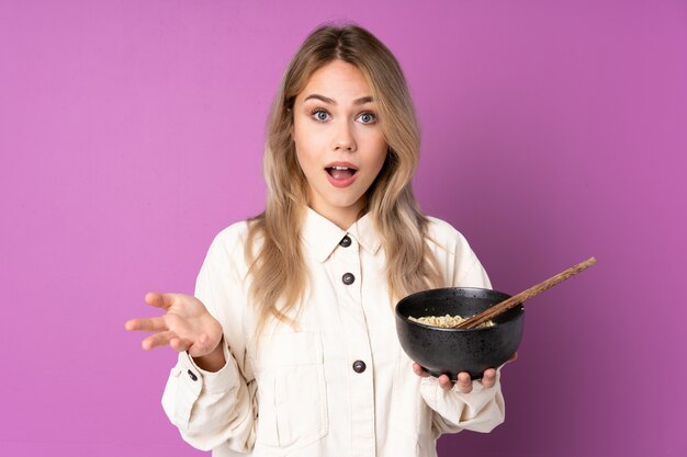 Teenager girl on purple wall with shocked facial expression while holding a bowl of noodles with chopsticks