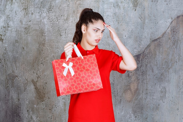 Teenager girl in red shirt holding a red shopping bag and looks confused and thoughtful