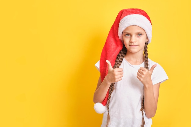 Teenager girl in santa claus hat showing thumbs up gesture, yellow background, copy space