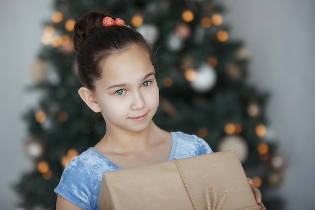 Teenager girl sits near a Christmas tree with a gift box in her hands. New Year. A pleasant surprise.