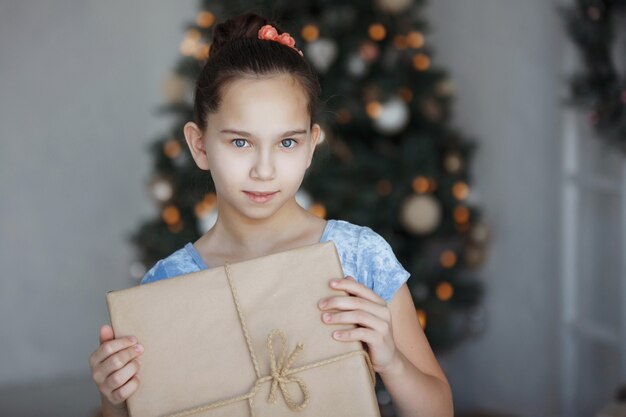 Photo teenager girl sits near a christmas tree with a gift box in her hands. new year. a pleasant surprise.