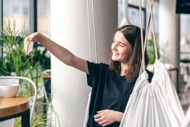 A teenager girl sits in a suspended hammock and takes a selfie on a smartphone