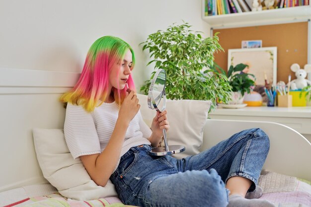 Teenager girl sitting at home on bed with makeup mirror painting her lips with lip gloss