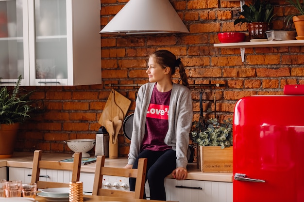 Teenager girl sitting at kitchen. Loft style kitchen with brick walls and red fridge.