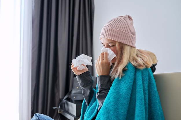 Teenager girl sneezing in handkerchief, female with symptoms of illness sitting at home covered with warm blanket and wearing knitted hat