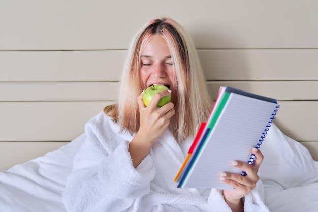 Foto studentessa adolescente con mela verde e quaderno scolastico giovane femmina impara a casa seduta a letto mangiando frutti naturali