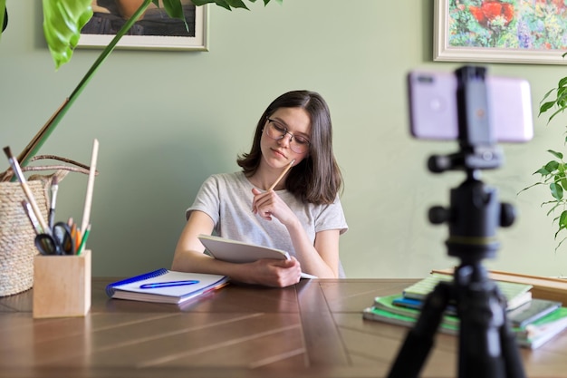 Teenager girl studying online using smartphone, female sitting at home at table with school books, listening talking at video conference. Distance learning, e-learning, technology, education, teens