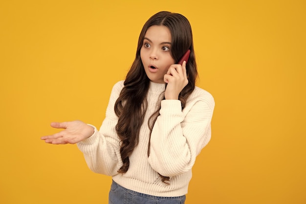 10代の少女はスマートフォンを使用して携帯電話でチャットするソーシャルメディアを使用する黄色の背景の上に分離されたスタイリッシュでカジュアルな流行の服を着る驚いた10代の少女