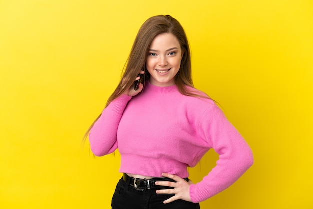 Teenager girl using mobile phone over isolated yellow background posing with arms at hip and smiling