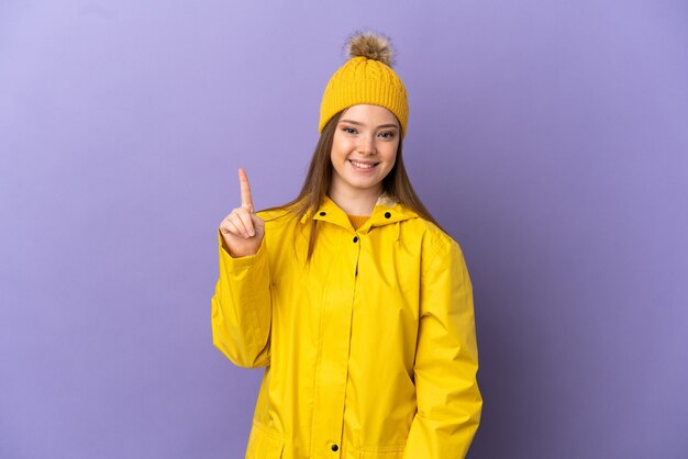 Девушка-подросток в непромокаемом пальто на изолированном фиолетовом фоне показывает и поднимает палец в знак лучших