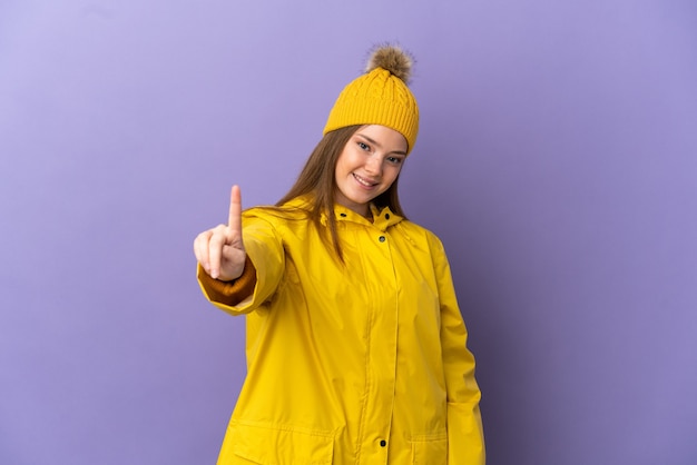 Teenager girl wearing a rainproof coat over isolated purple background showing and lifting a finger