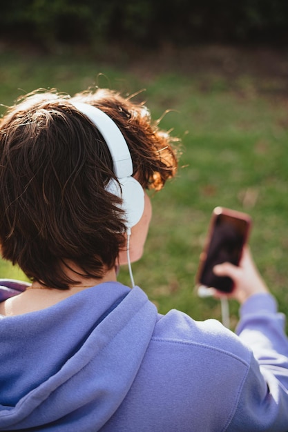 A teenager girl wearing white headphones to listen to music while sitting in the grass. She is using a smart phone.