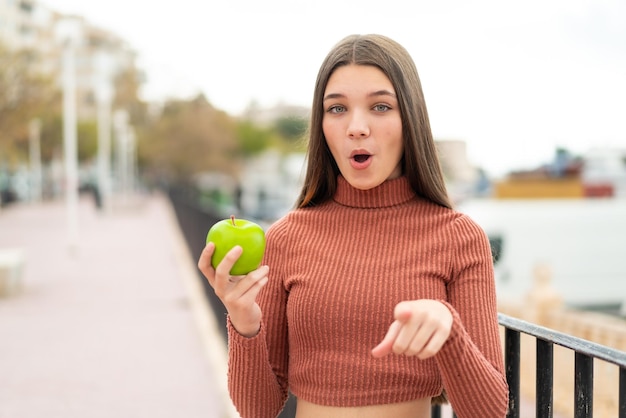 屋外でリンゴを持つティーンエイジャーの女の子が驚いて前を指している