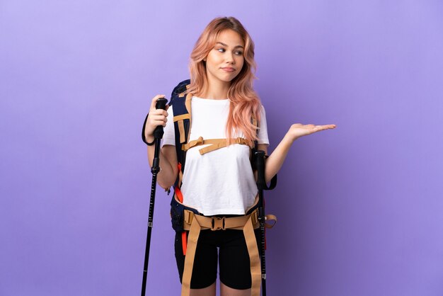 Teenager girl with backpack and trekking poles over isolated purple background holding copyspace with doubts