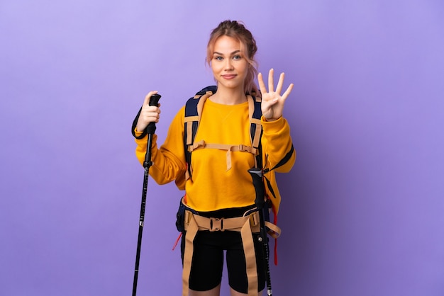 Девушка-подросток с рюкзаком и треккинговыми палками над изолированным фиолетовым счастьем и считает четыре пальцами