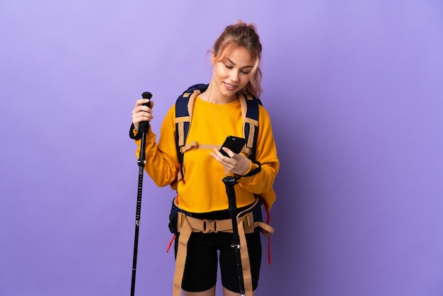 Teenager girl with backpack and trekking poles over isolated purple sending a message with the mobile