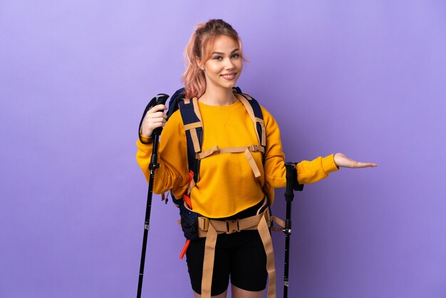 Teenager girl with backpack and trekking poles over isolated purple wall extending hands to the side for inviting to come