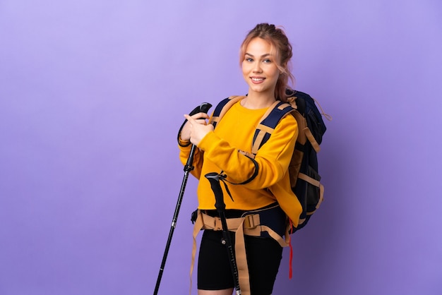 Teenager girl with backpack and trekking poles over isolated purple wall pointing back