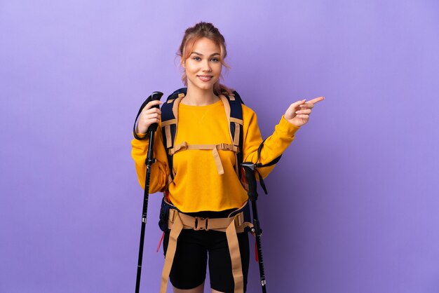 Teenager girl with backpack and trekking poles over isolated purple wall pointing finger to the side