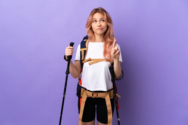 Teenager girl with backpack and trekking poles over isolated purple with fingers crossing and wishing the best