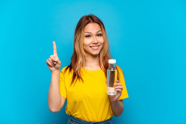 Teenager girl with a bottle of water over isolated blue background showing and lifting a finger in sign of the best