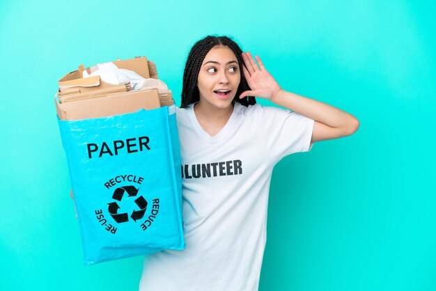 Teenager girl with braids holding a bag to recycle listening to something by putting hand on the ear