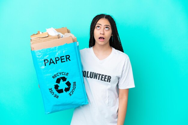 Teenager girl with braids holding a bag to recycle looking up and with surprised expression