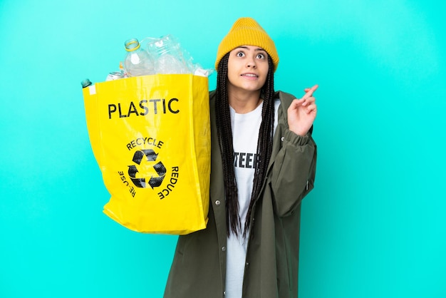 Teenager girl with braids holding a bag to recycle with fingers crossing and wishing the best