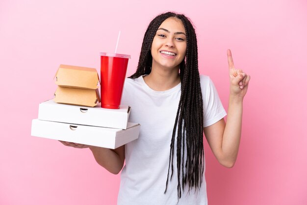 Ragazza adolescente con le trecce che tengono pizze e hamburger su sfondo rosa isolato che indica una grande idea