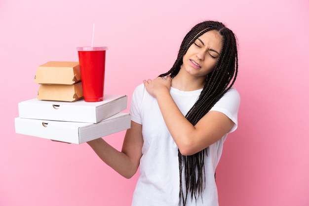 Девушка-подросток с косами, держащая пиццу и гамбургеры на изолированном розовом фоне, страдает от боли в плече за то, что приложила усилия