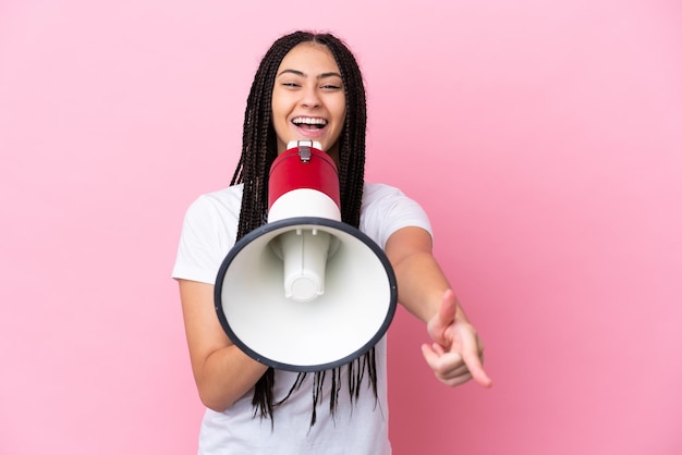 Teenager girl with braids over isolated pink background shouting through a megaphone to announce something while pointing to the front