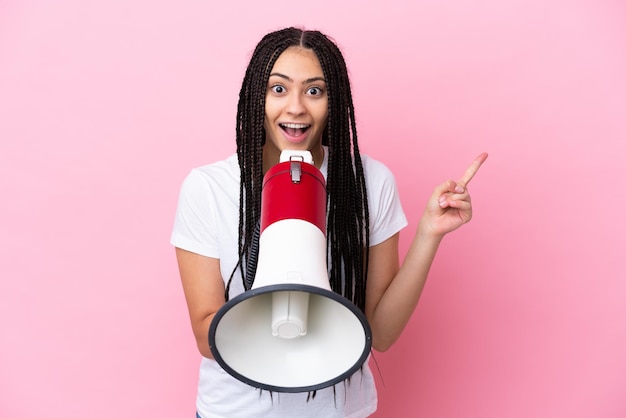 Teenager girl with braids over isolated pink background shouting through a megaphone and pointing side