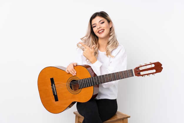 Teenager girl with guitar over isolated white  pointing to the side to present a product