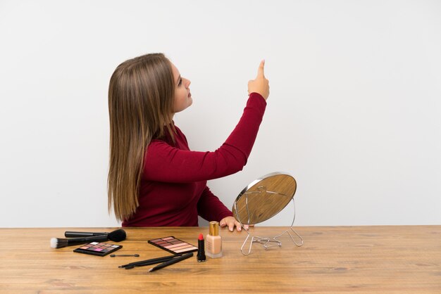 Teenager girl with makeup palette and cosmetics in a table pointing back with the index finger