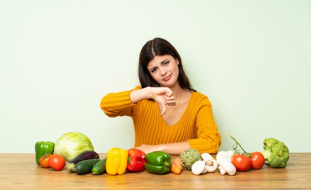 Teenager girl with many vegetables showing thumb down with negative expression