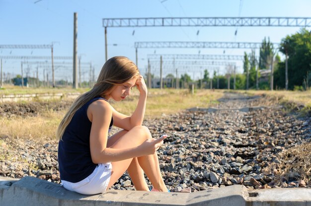 未完成のレールトラックに座っている携帯電話を持つティーンエイジャーの少女